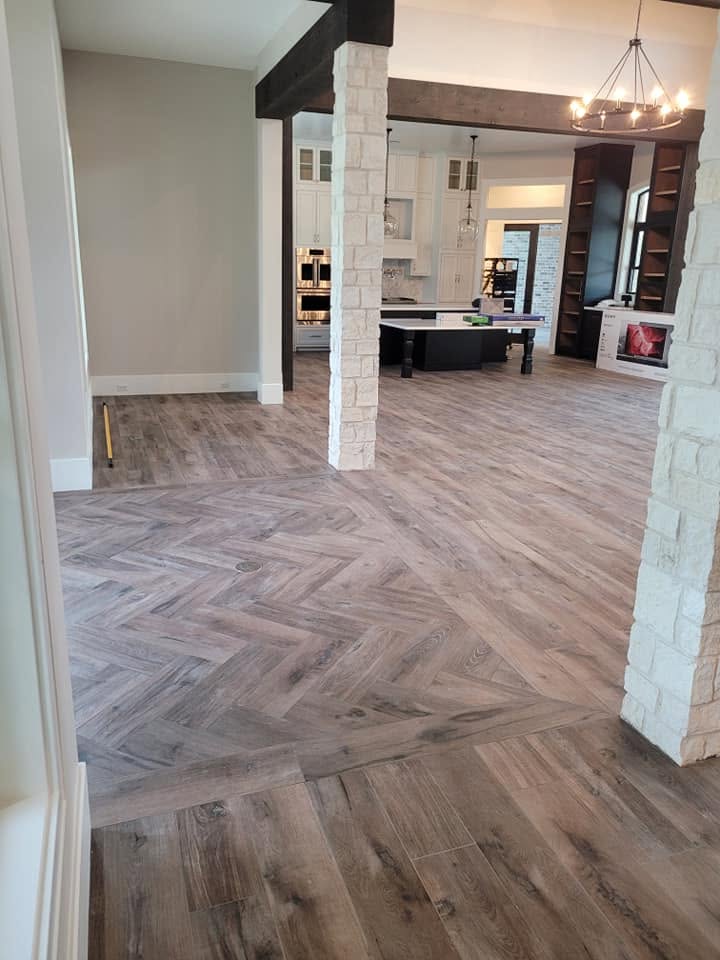 Open-concept home interior with herringbone wood flooring, white support columns, and modern kitchen visible in background